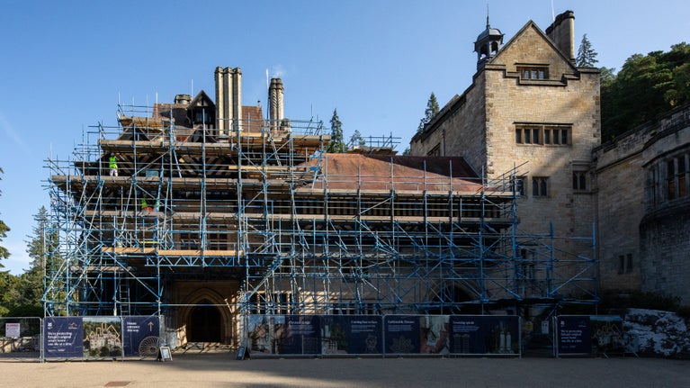 Cragside's main entrance is surrounded by extensive scaffolding during roof conservation work. Workers are visible on the scaffold platforms, and protective hoardings line the front of the building.